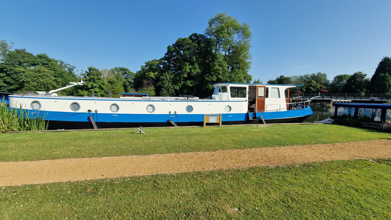 Che Bella Vita, a blue and white Dutch barge, moored on the Thames beside a grassy bank on a bright summer day