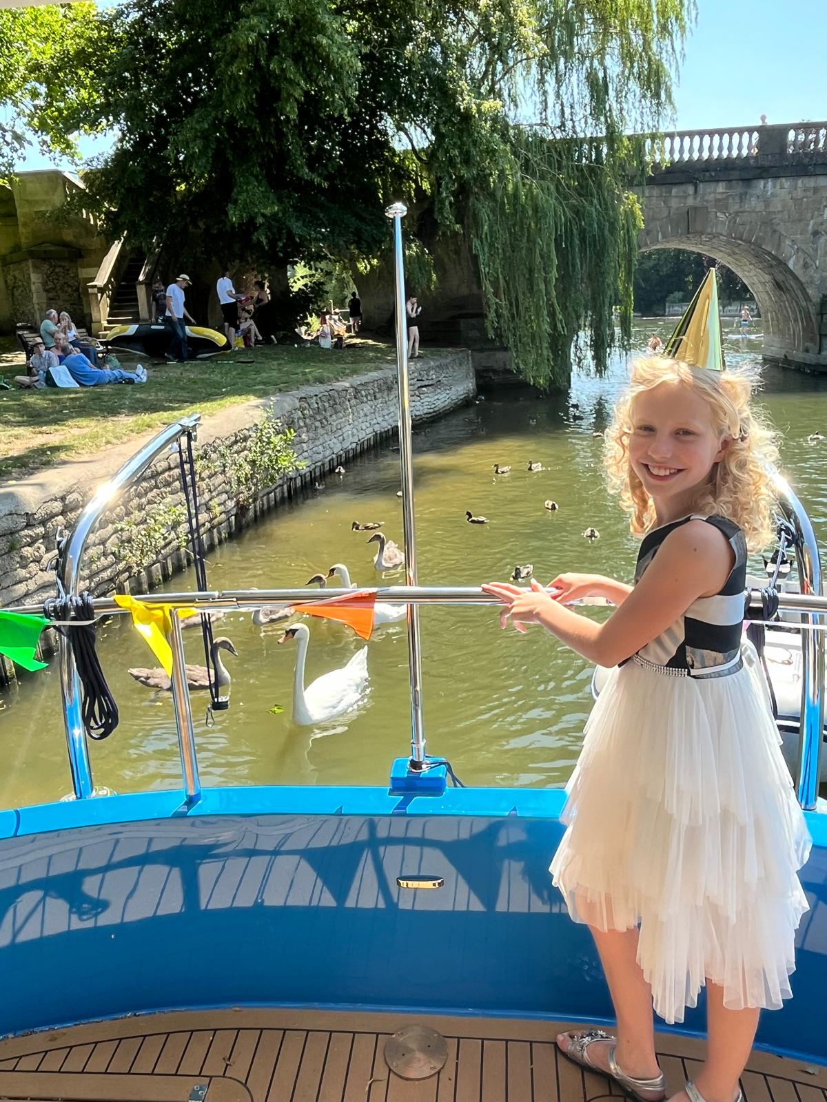 A young girl in a party hat at Che Bella Vita's rail, with swans on the river and a stone bridge behind