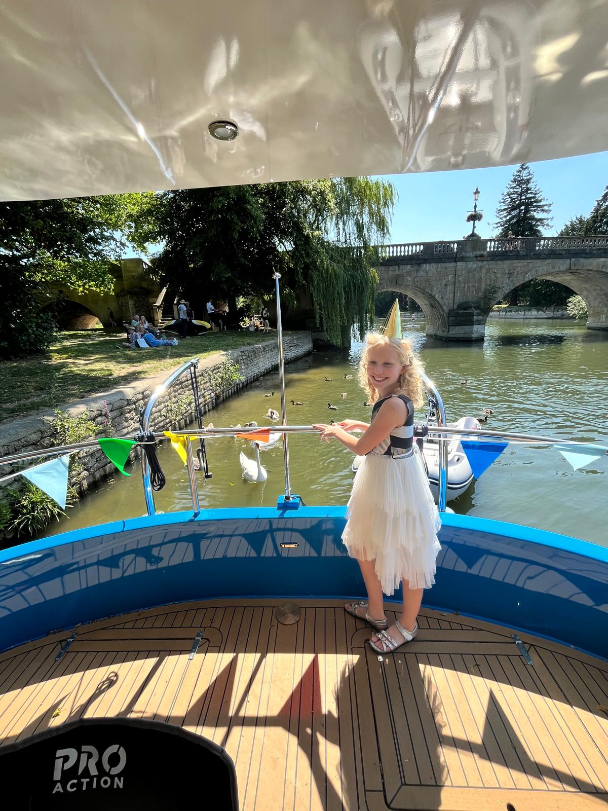 A young girl in a party hat on Che Bella Vita's back deck, with bunting on the rail, swans on the river and a stone bridge beyond